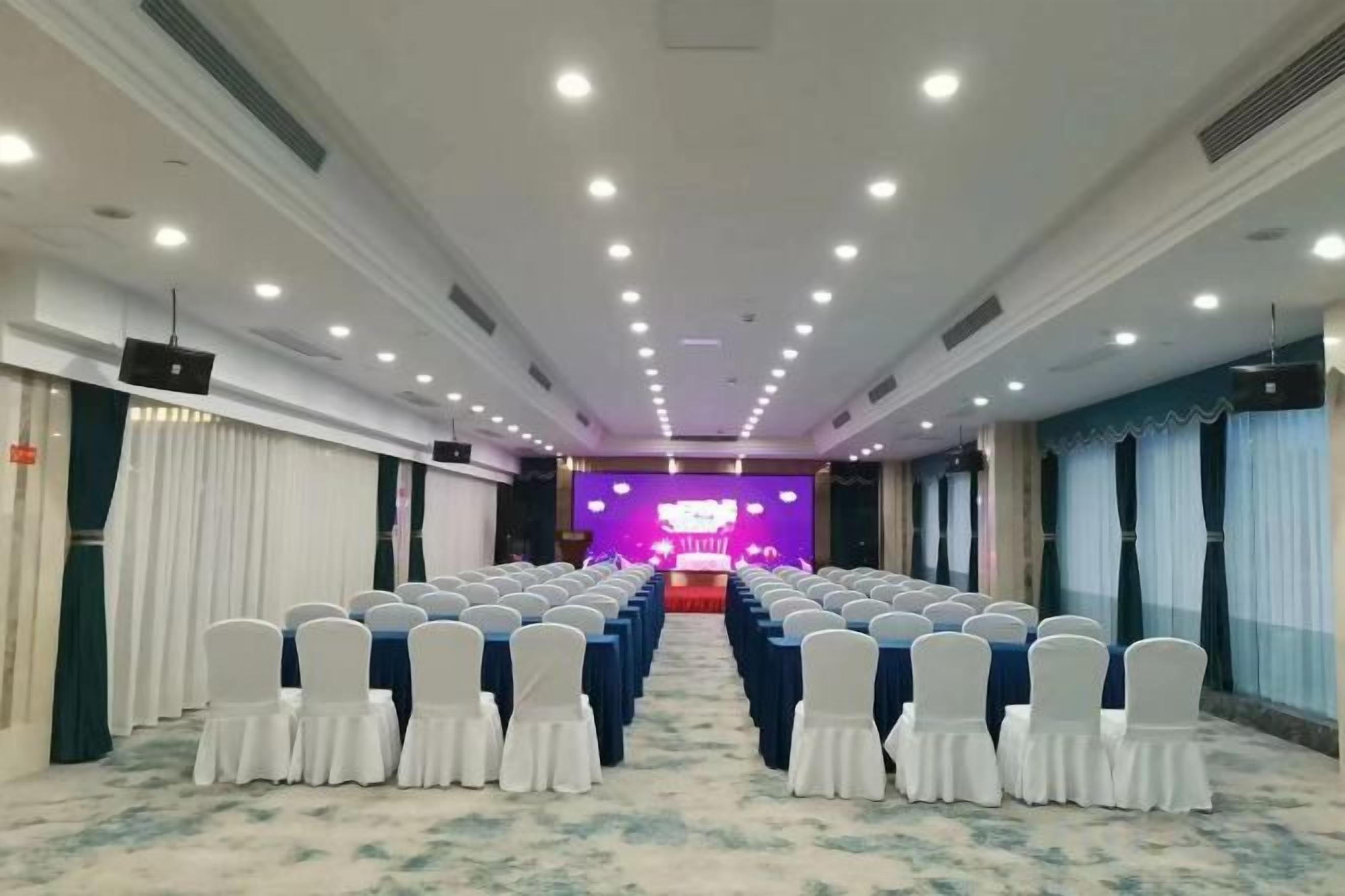 A conference room with rows of chairs, a stage with a purple backdrop, and a carpeted floor.