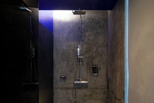 Un área de ducha moderna con un cabezal de ducha tipo lluvia vertical y otro cabezal de ducha manual, frente a una pared texturizada.