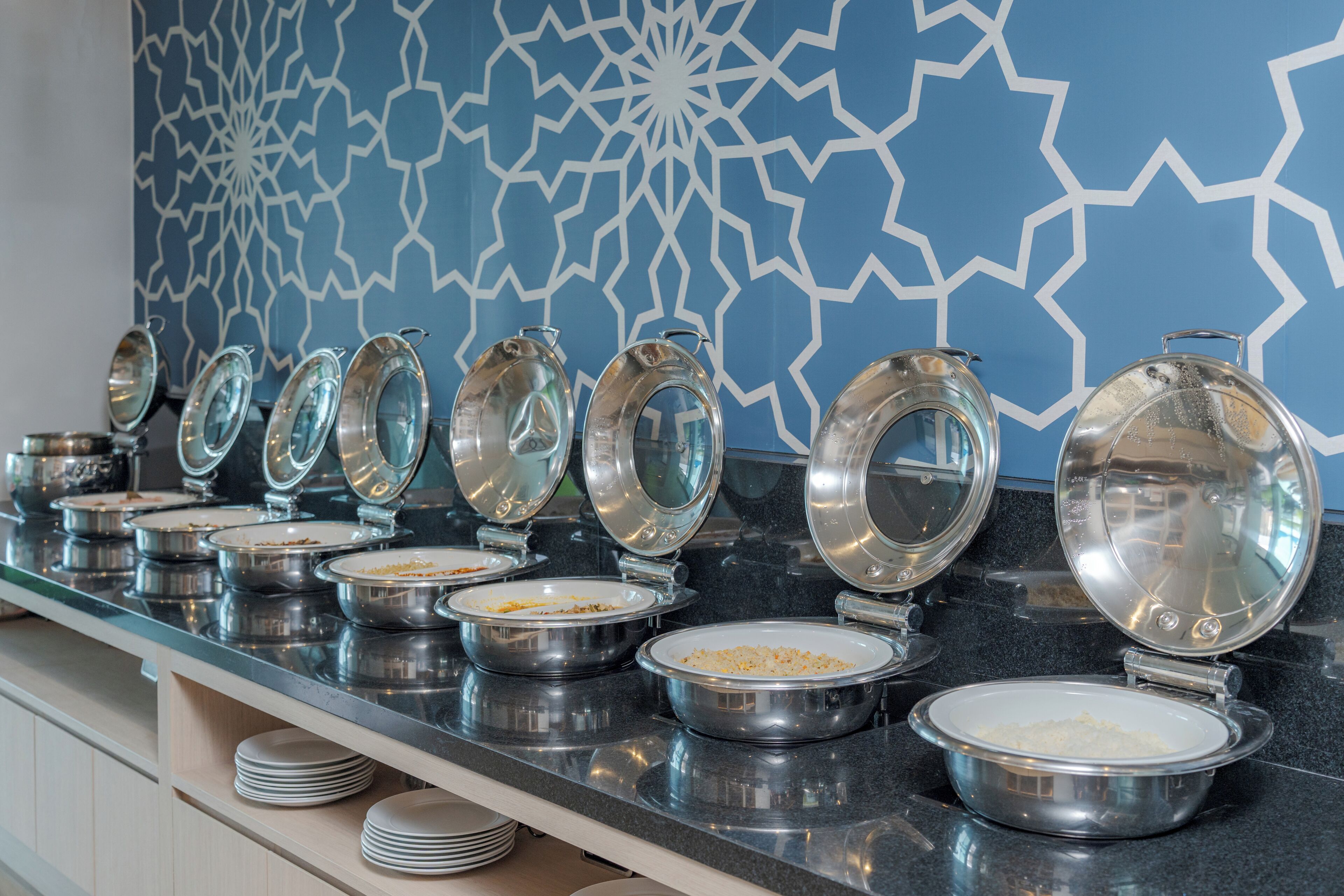 A buffet line with multiple bowls of food, each with a lid, on a counter with a blue patterned wall in the background.