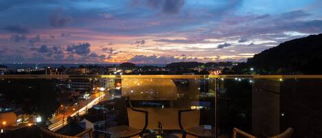 A balcony with wicker chairs and a table, overlooking a cityscape at dusk with visible street lights.