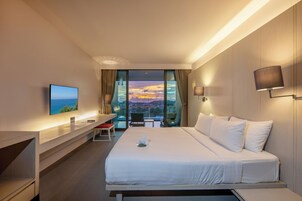 A hotel room with a bed, a desk, a chair, a TV, and a view of the sunset through large windows.