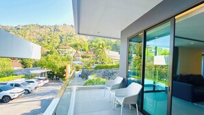 Un balcón con puerta de cristal, zona de estar con sillas, piscina y vista a una calle y edificios al fondo.