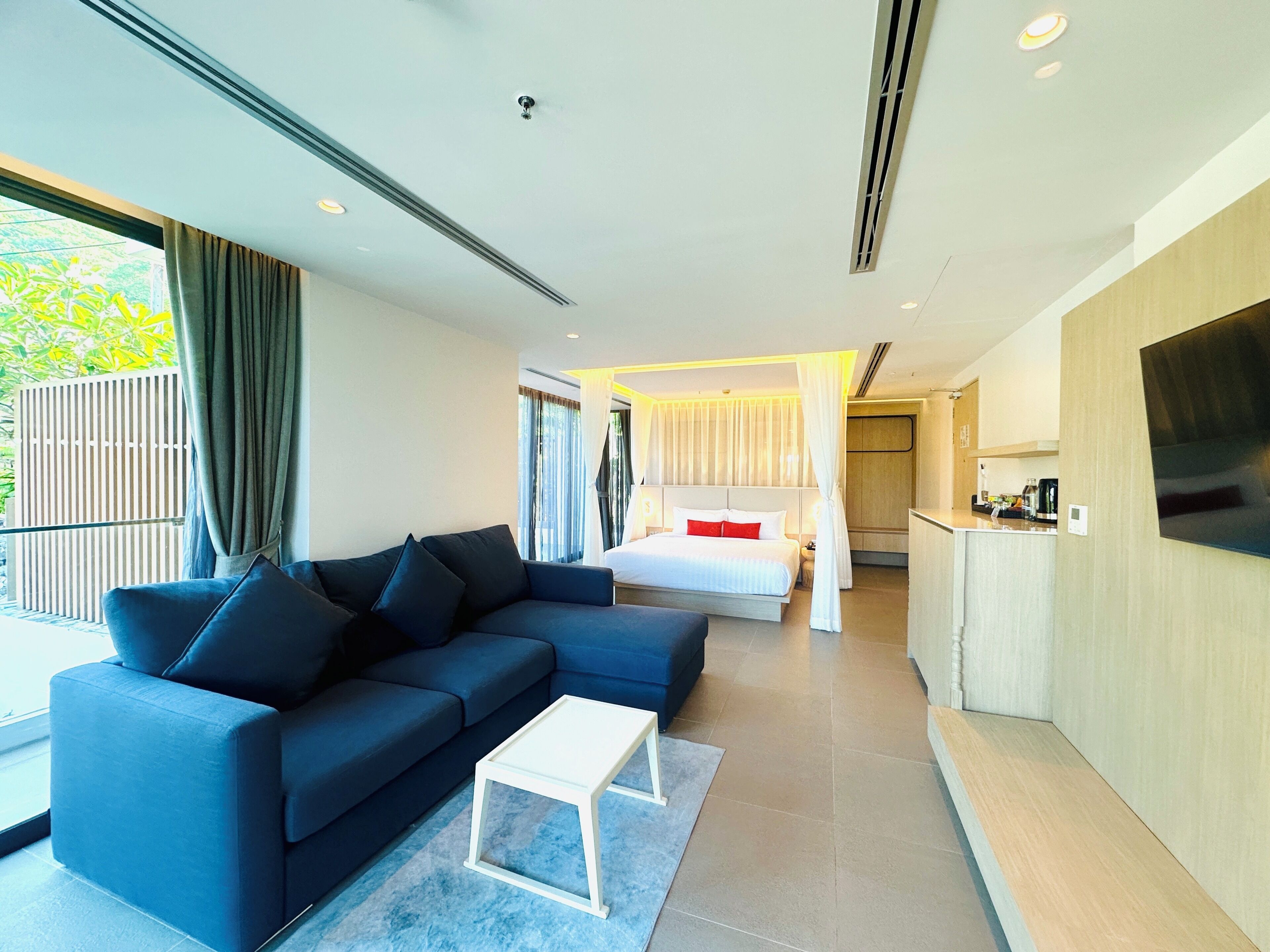 Habitación de hotel moderna con sofá, una pequeña mesa blanca, cama con ropa de cama blanca y almohadas rojas, televisión y balcón con cortinas.