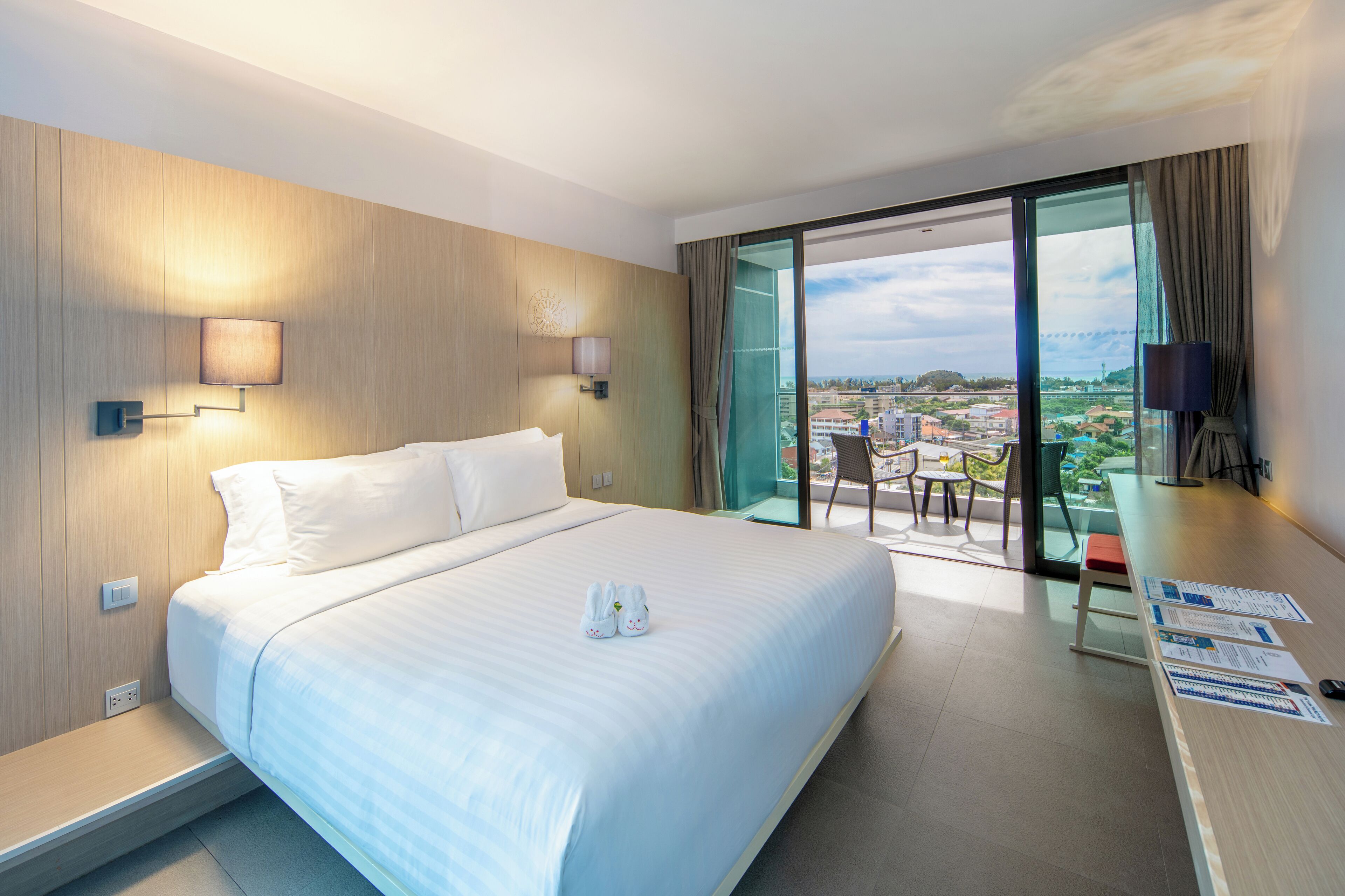 A hotel room with a large bed, a desk with a chair, and a balcony with a city view.
