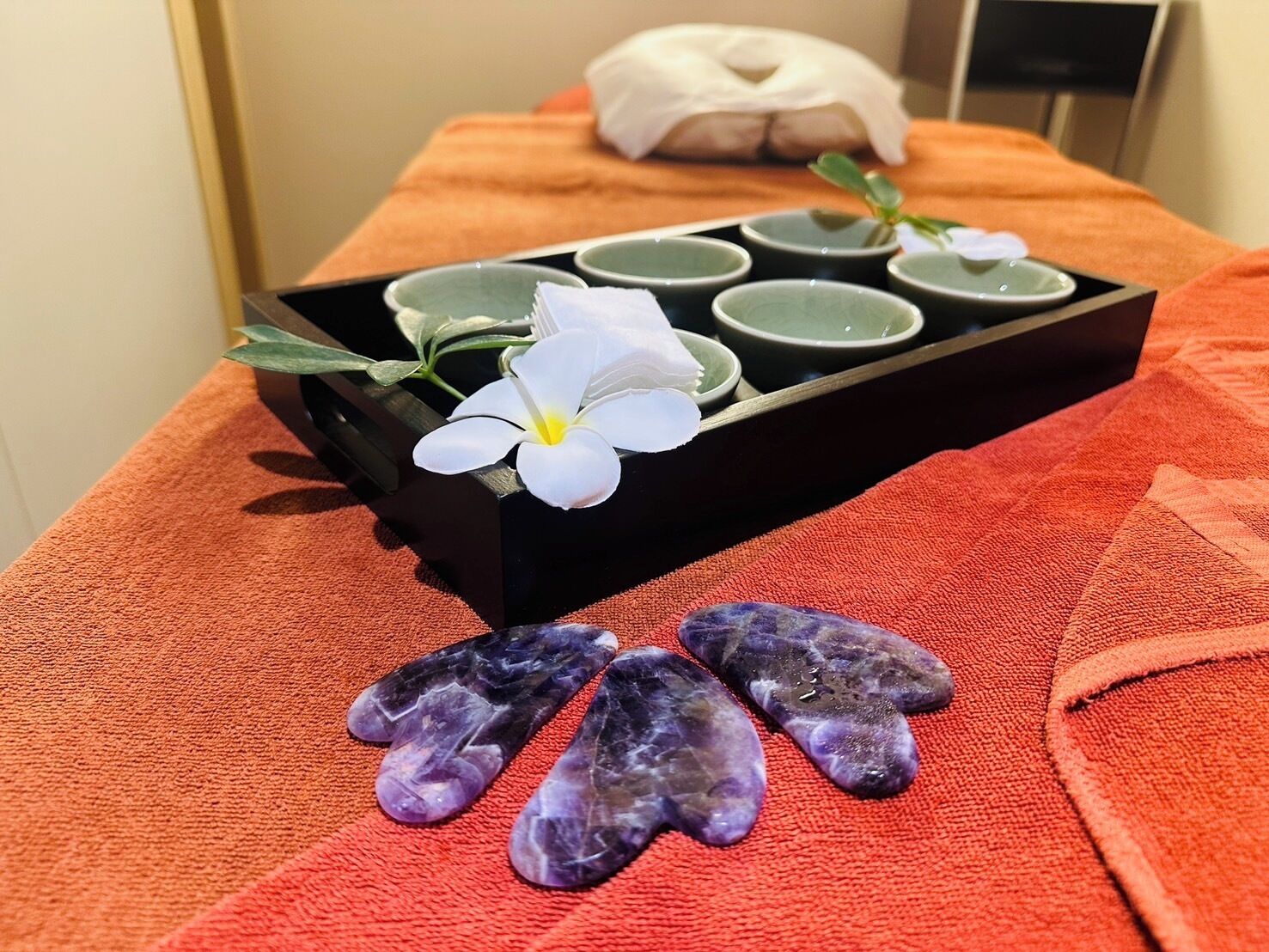 A massage table with a tray of green cups, a white flower, and purple stones.