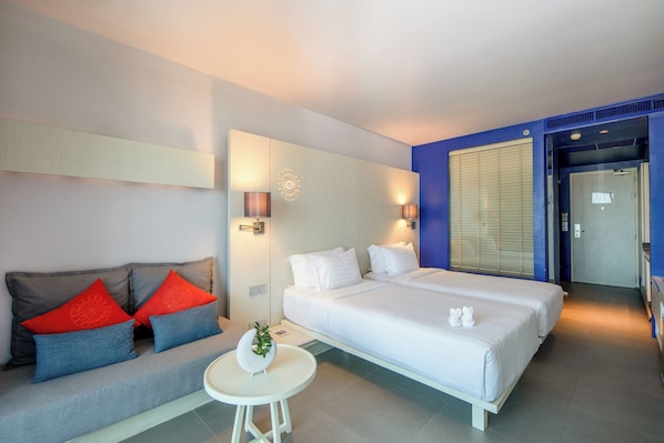 Una habitación de hotel moderna con una cama, un sofá con cojines rojos y azules, una mesita redonda y una lámpara fijada a la pared.