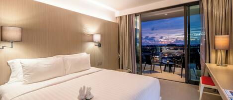 Habitación de hotel con una cama grande, un balcón y vistas a la ciudad, y un escritorio con una silla.