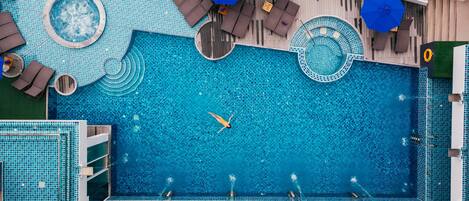 En vy från ovan över en stor swimmingpool med flera bubbelpooler, avkopplingsområden och en bar vid poolkanten.