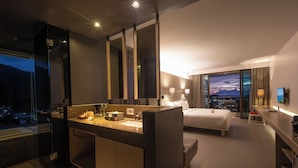 Habitación de hotel moderna con una cama grande, baño separado con tocador, escritorio y un ventanal con vistas a la ciudad.