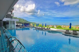 A swimming pool with multiple lanes, sun loungers, and umbrellas, surrounded by a modern building and a view of a hilly landscape.