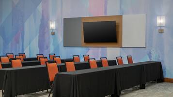 Sala de conferência com uma longa mesa, cadeiras, uma tela de TV e luminárias fixadas na parede.