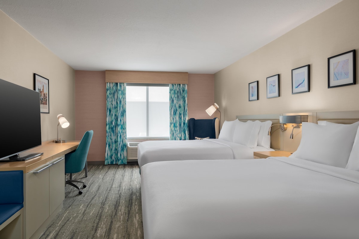 A hotel room with two beds, a desk, a chair, a TV, and a window with curtains.