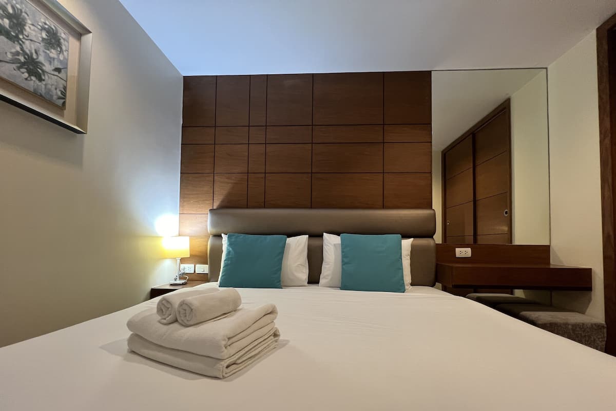 A hotel room with a large bed, a headboard with a wooden panel design, two pillows, a nightstand with a lamp, and a folded towel on the bed.