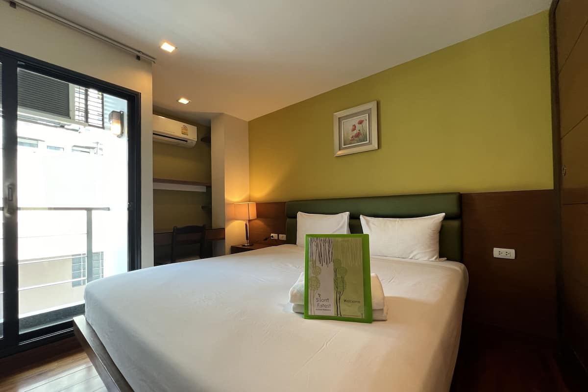 A hotel room with a large bed, a bedside table with a lamp, a framed picture on the wall, and an air conditioning unit.