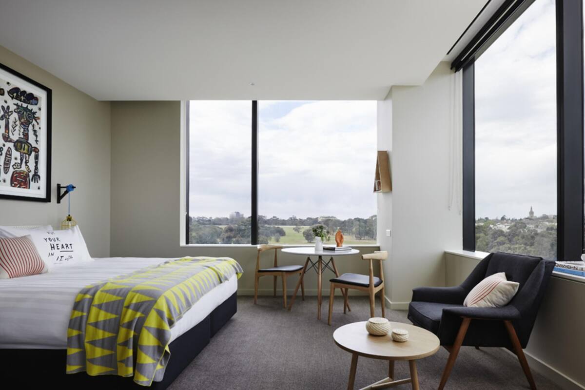 A modern hotel room with a large bed, a desk, two chairs, and a view of the cityscape.