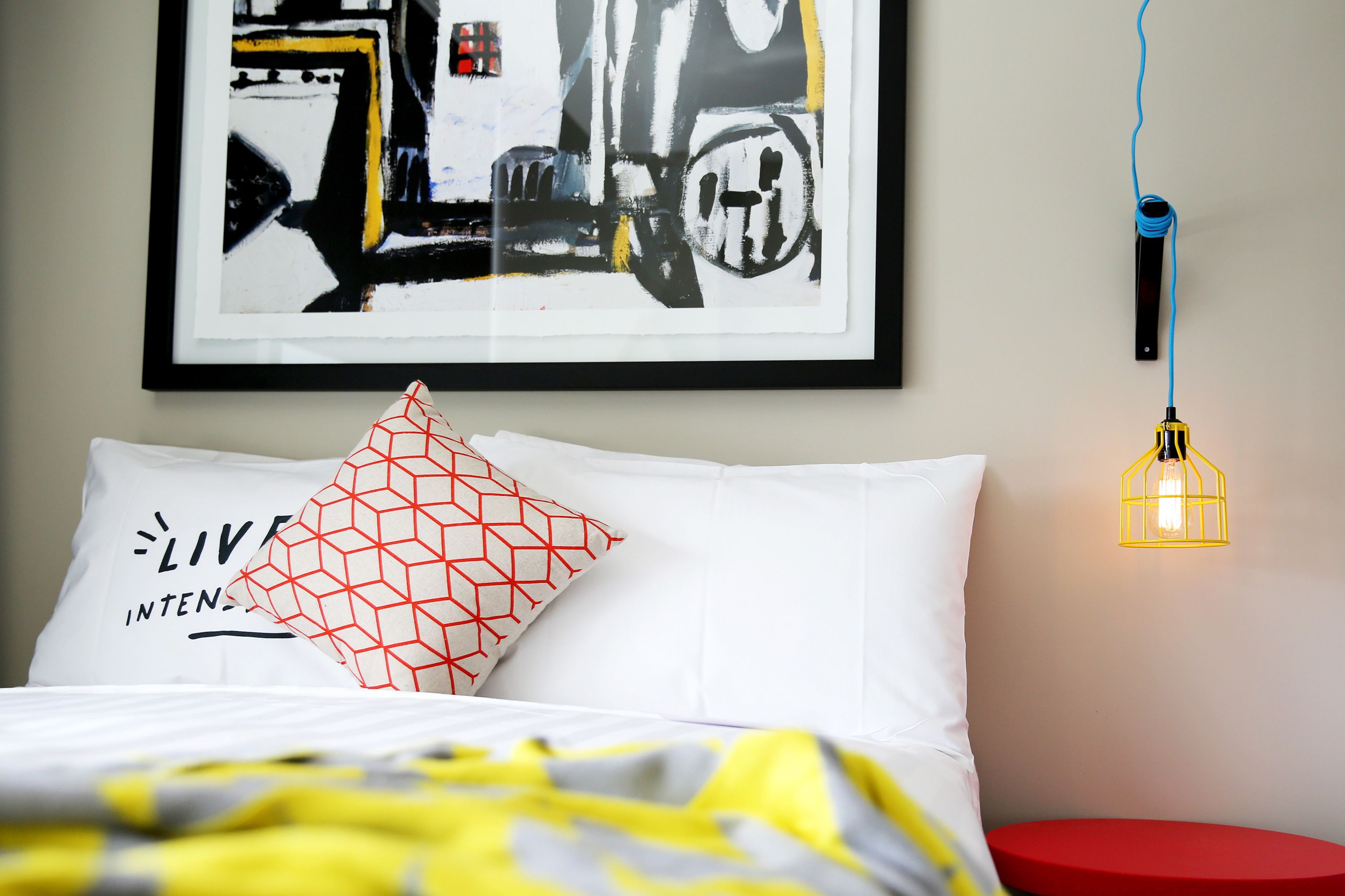 A bed with a white pillow and a red geometric pillow, a yellow and gray blanket, a red stool, and a framed abstract painting on the wall.