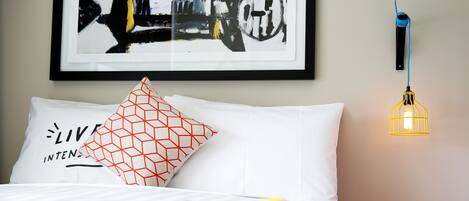 Una cama con almohada blanca y una almohada roja con diseño geométrico, una manta amarilla y gris, un taburete rojo y un cuadro abstracto enmarcado en la pared.