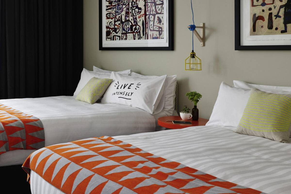 A hotel room with two beds, a nightstand, a wall-mounted light fixture, and a framed abstract painting.