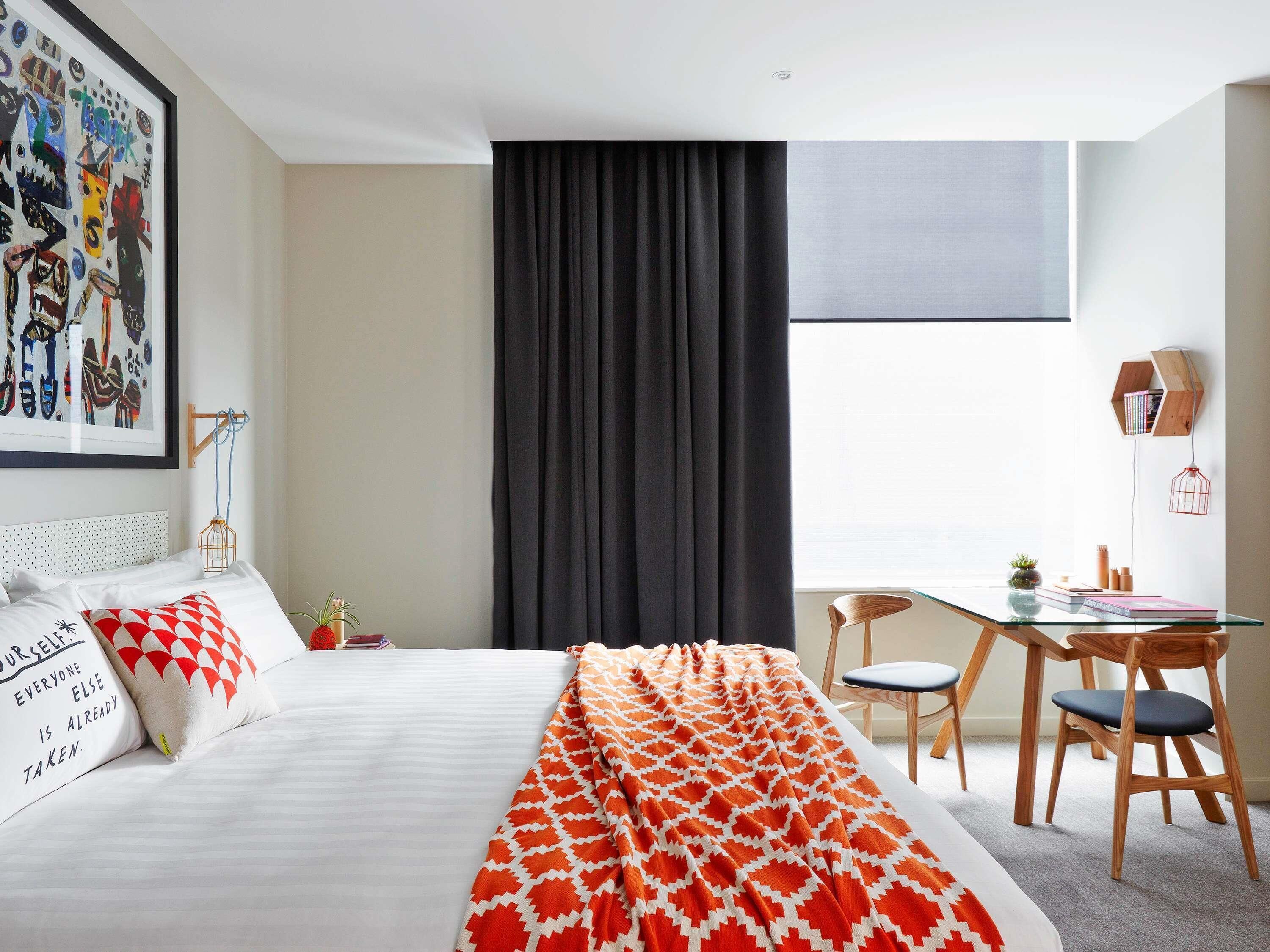 A hotel room with a bed, a desk, a chair, a window with curtains, and a wall-mounted shelf with books.
