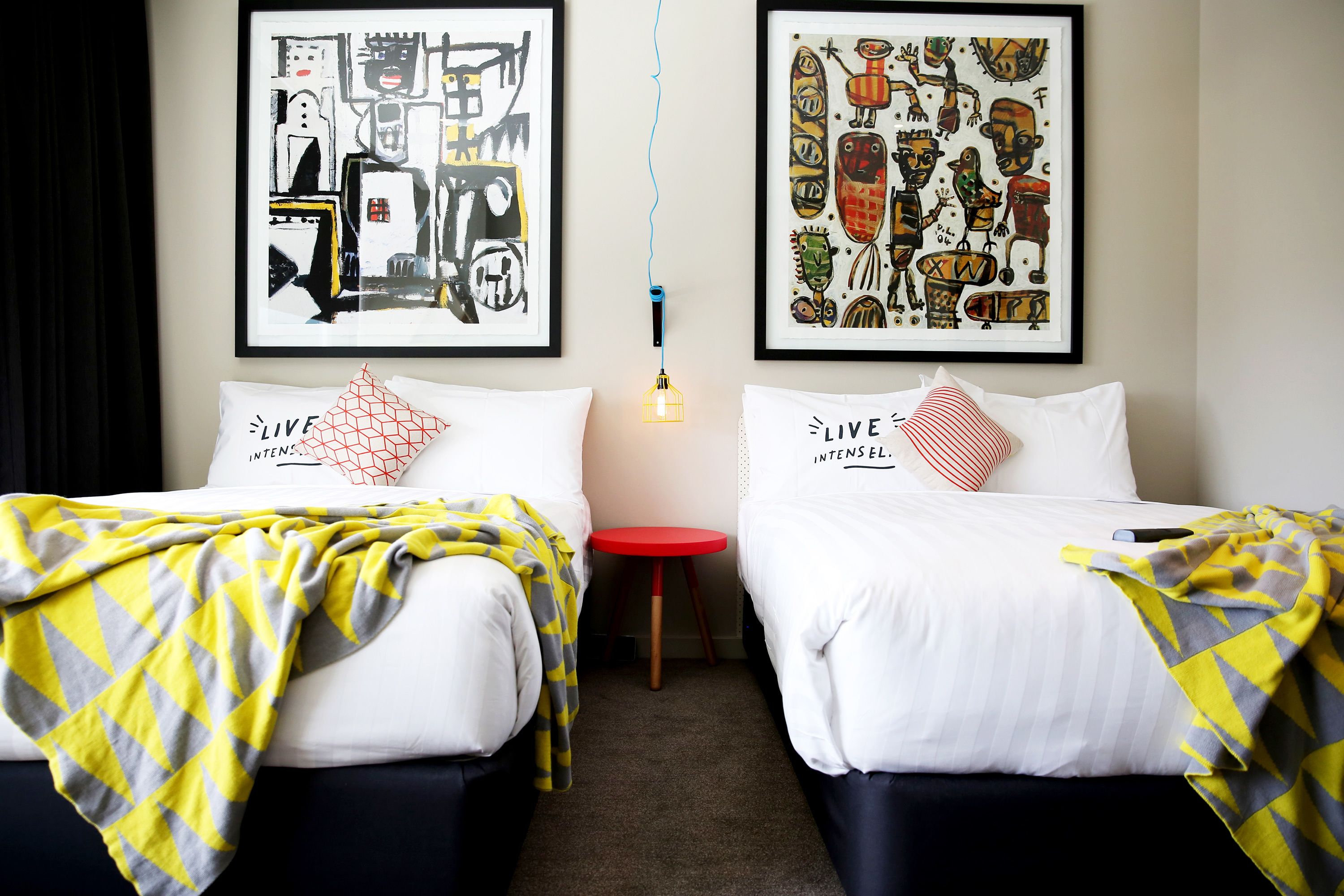 A hotel room with two beds, each with a white bedspread and a yellow and gray checkered blanket. There are two framed artworks on the wall, a red stool, and a hanging light fixture.