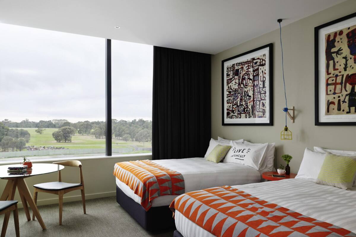 A hotel room with two beds, a small table, a chair, and a large window offering a view of the countryside.