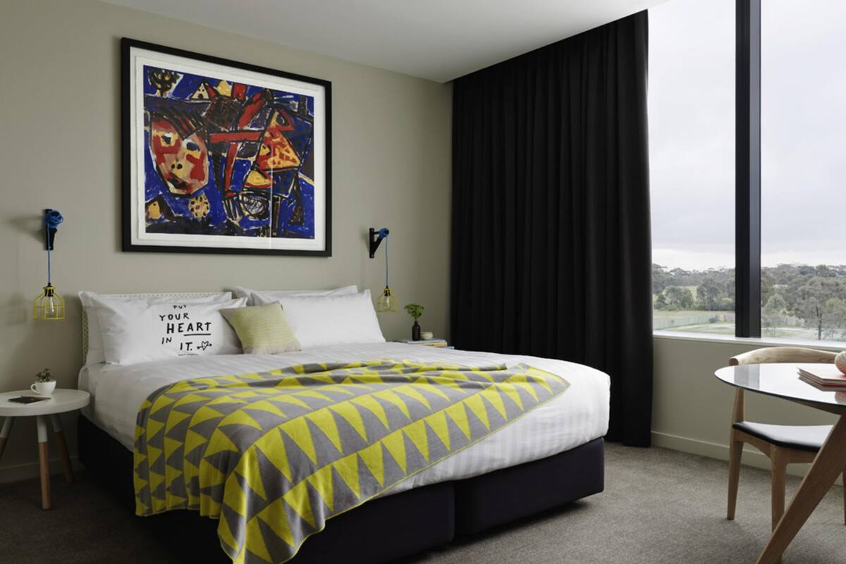 A hotel room with a bed, a painting on the wall, a window with curtains, and a view of the outdoors.