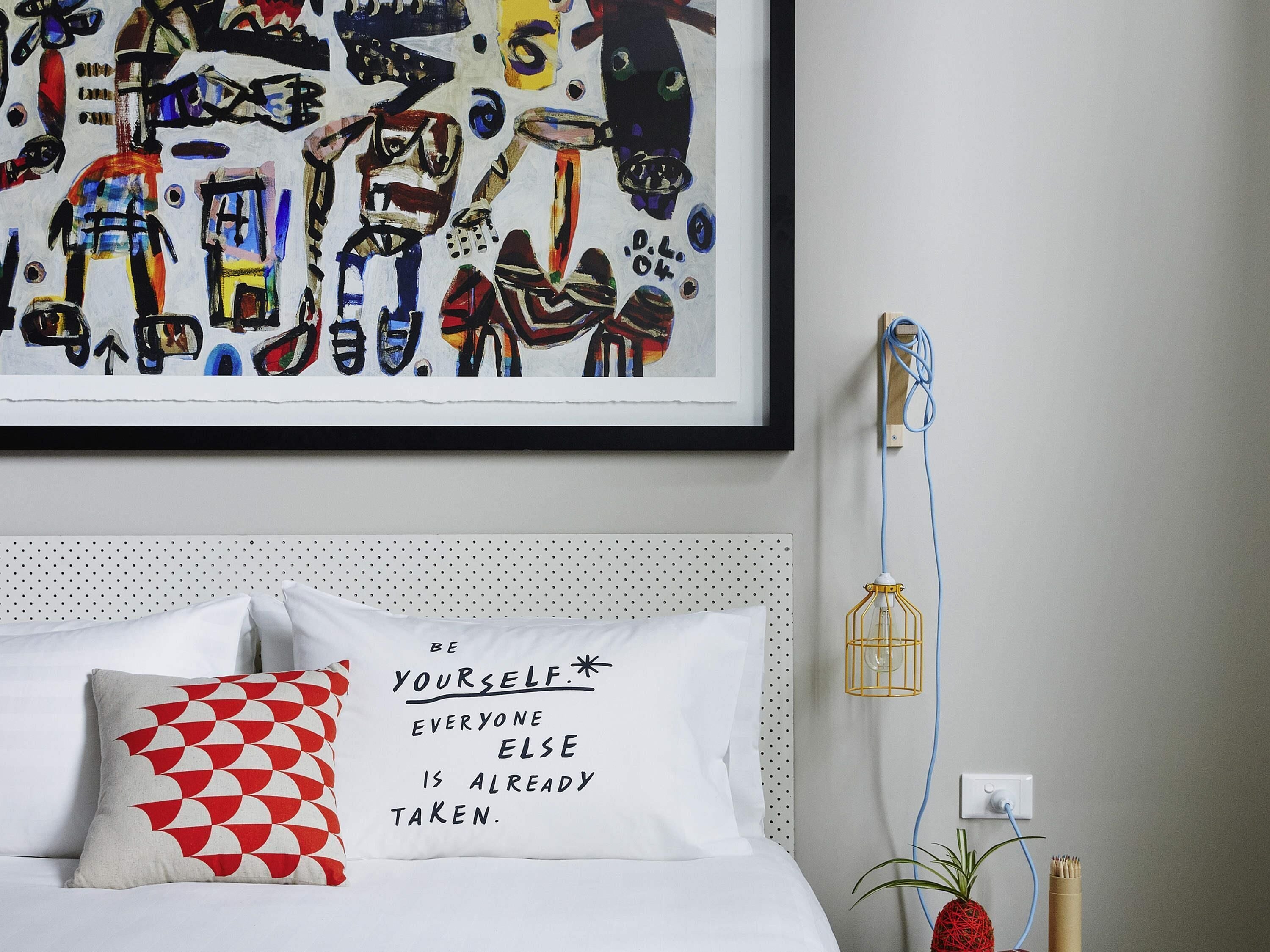 Un dormitorio con cama, un almohadón con una frase motivadora, una obra de arte en la pared, un enchufe y una planta.