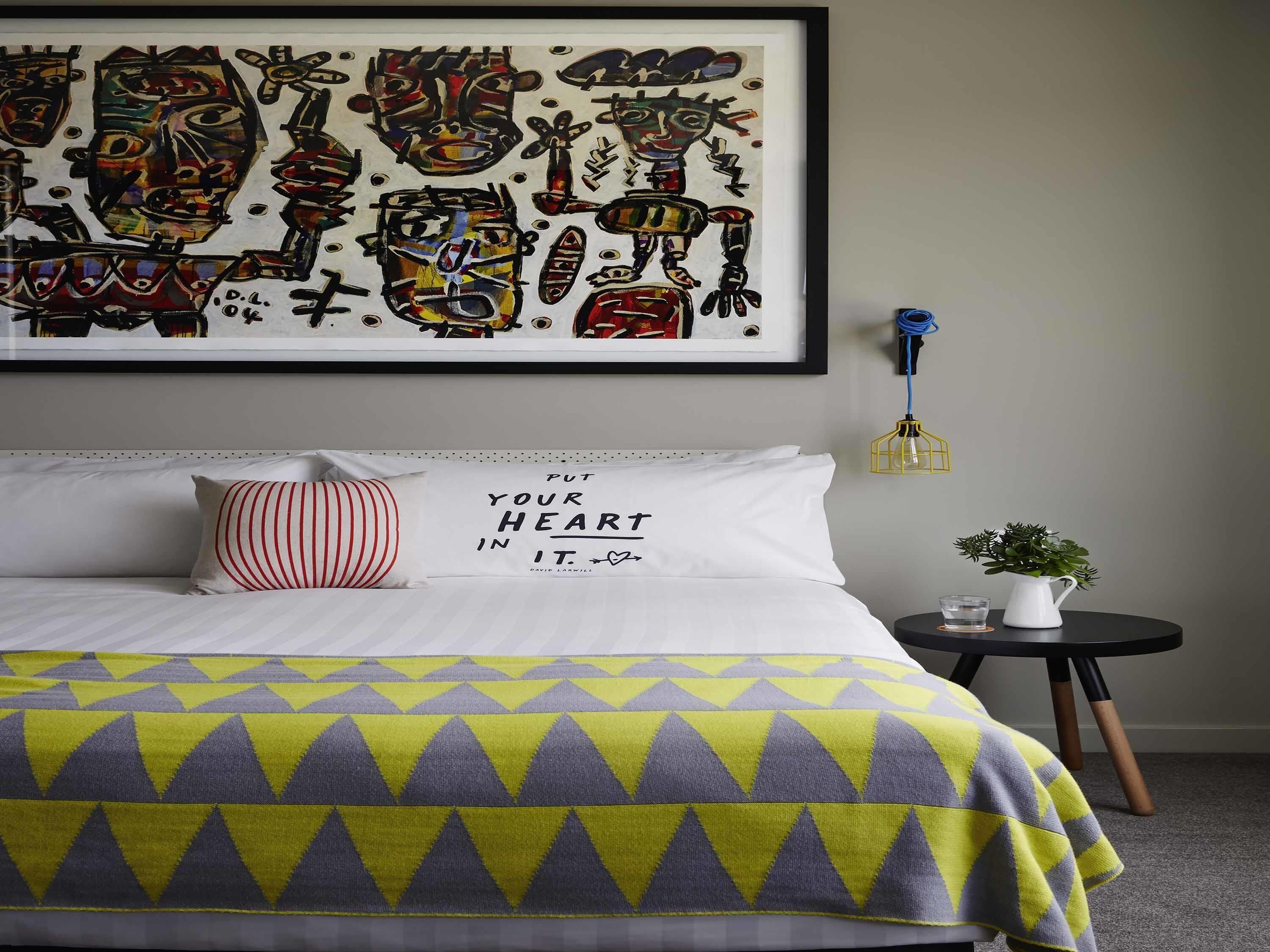 Une chambre à coucher avec un lit, un oreiller orné d’une citation, une œuvre d’art encadrée, une petite table avec une plante et un luminaire fixé au mur.