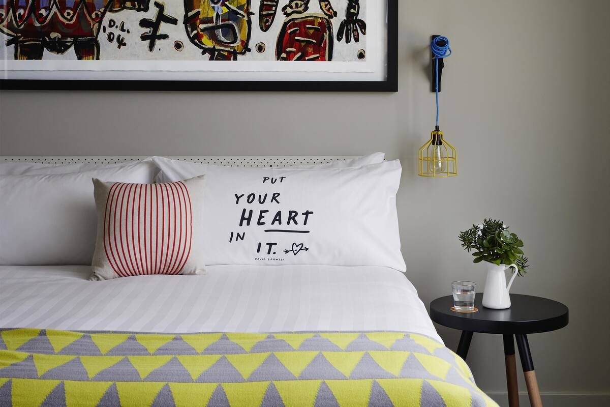A neatly made bed with a geometric-patterned bedspread, a pillow with a motivational quote, a framed artwork above the bed, a small table with a plant and a glass, and a wall-mounted light fixture.