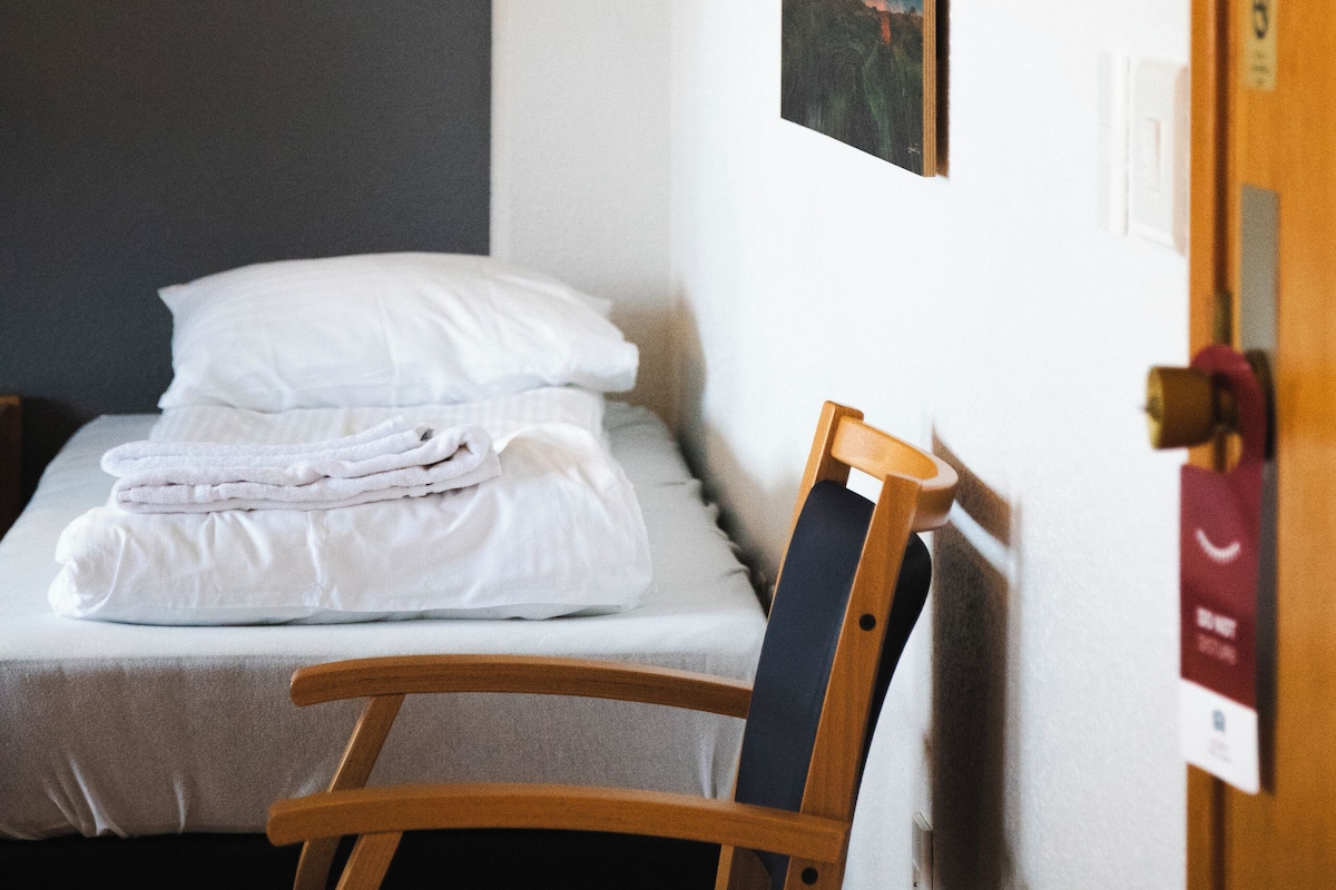 Una cama individual con sábanas blancas, una silla de madera, un cuadro de la aurora boreal y una puerta con un letrero rojo.