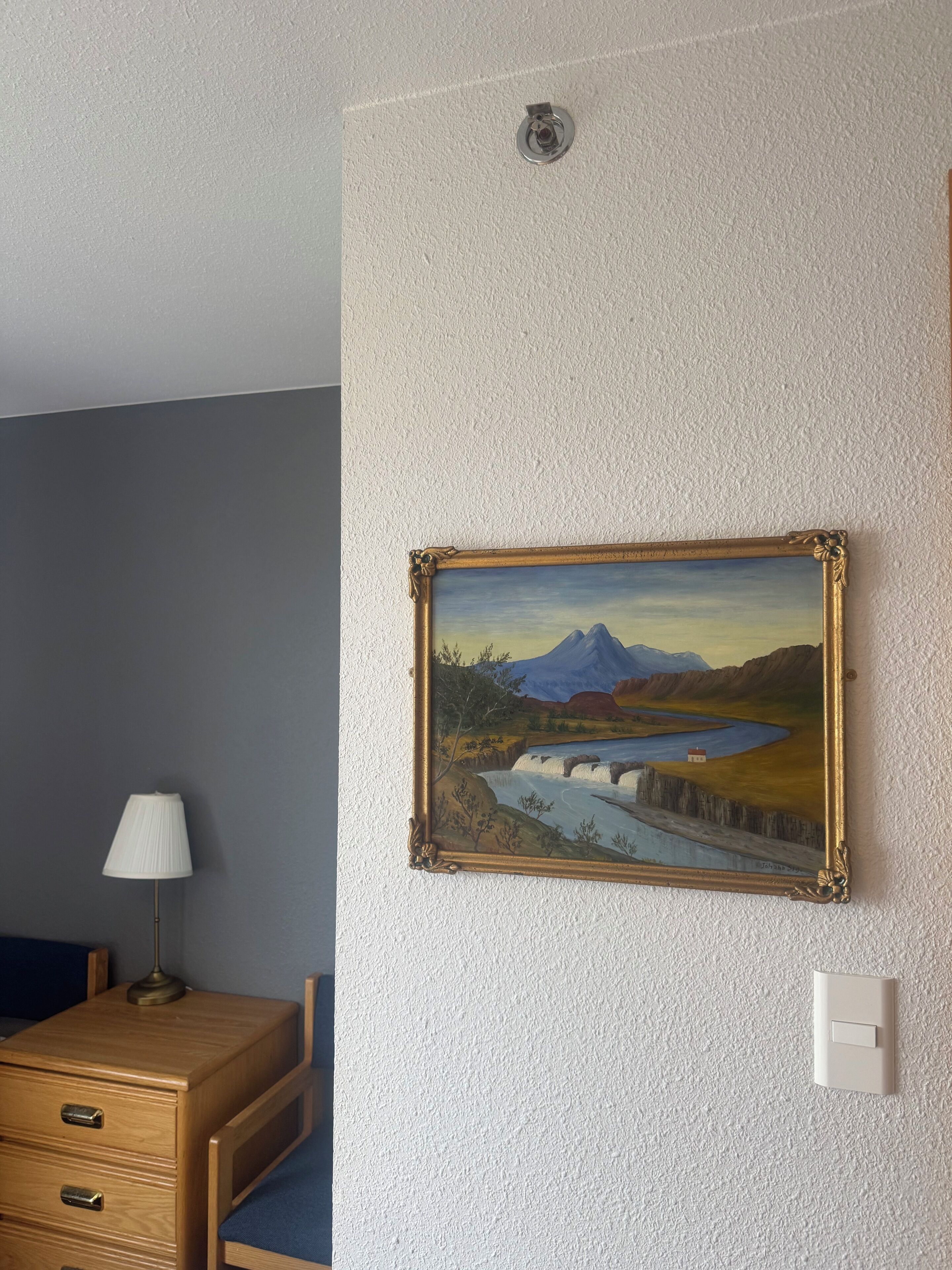 A room with a painting of a landscape, a lamp, a dresser, and a light switch on the wall.