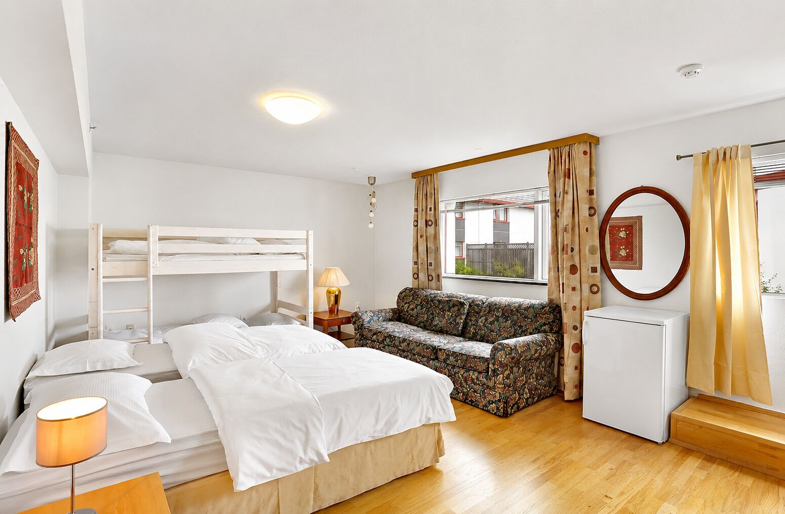 Ein Zimmer mit einem Etagenbett, einer Schlafcouch, einem kleinen Tisch mit Lampe, einem Heizkörper und einem fenster mit Vorhängen.