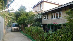 A narrow street with a parked car, a house with a green awning, and dense foliage. - Phurahong Homestay (Bangkok)