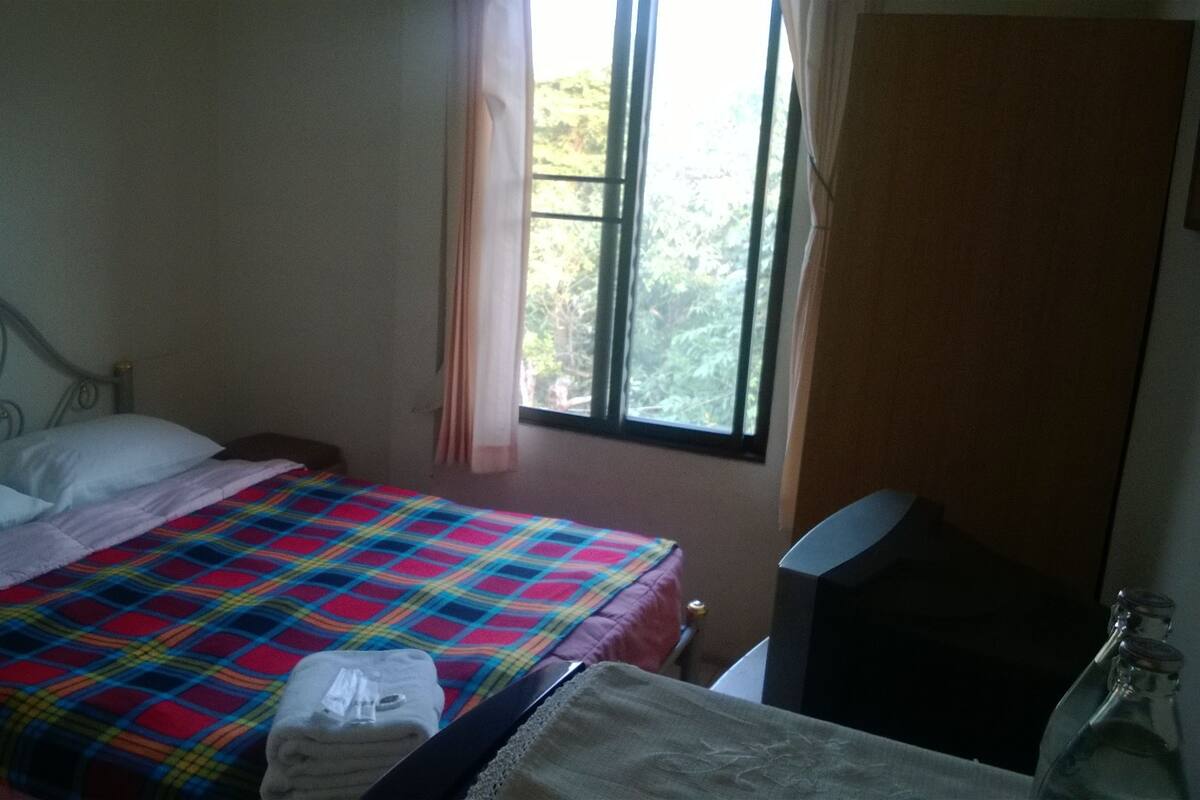 A bedroom with a metal bed frame, a colorful bedspread, a window with curtains, and a bedside table with a bottle.