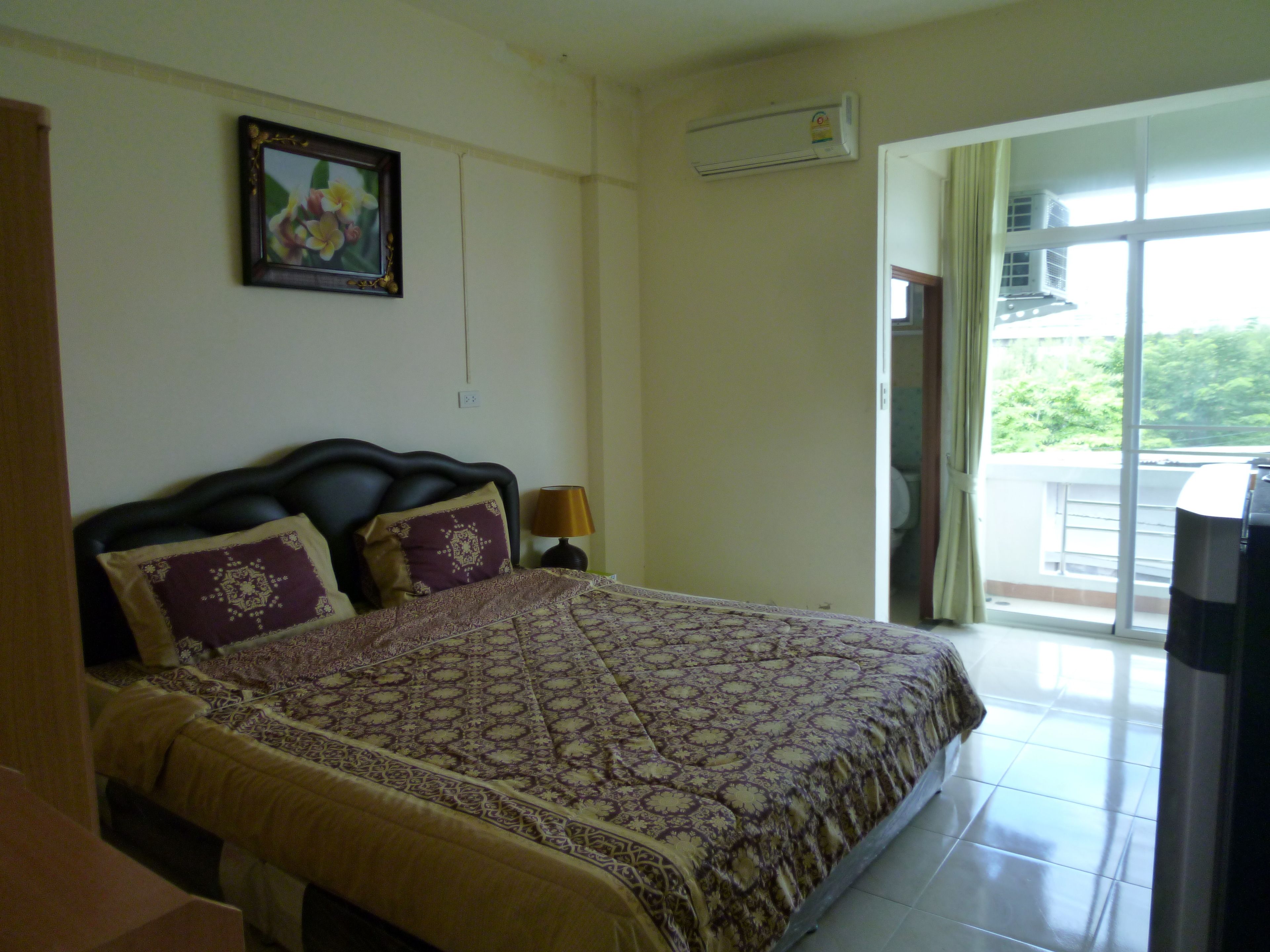 A bedroom with a bed, a nightstand, a lamp, a framed picture on the wall, and a window with a view of greenery.