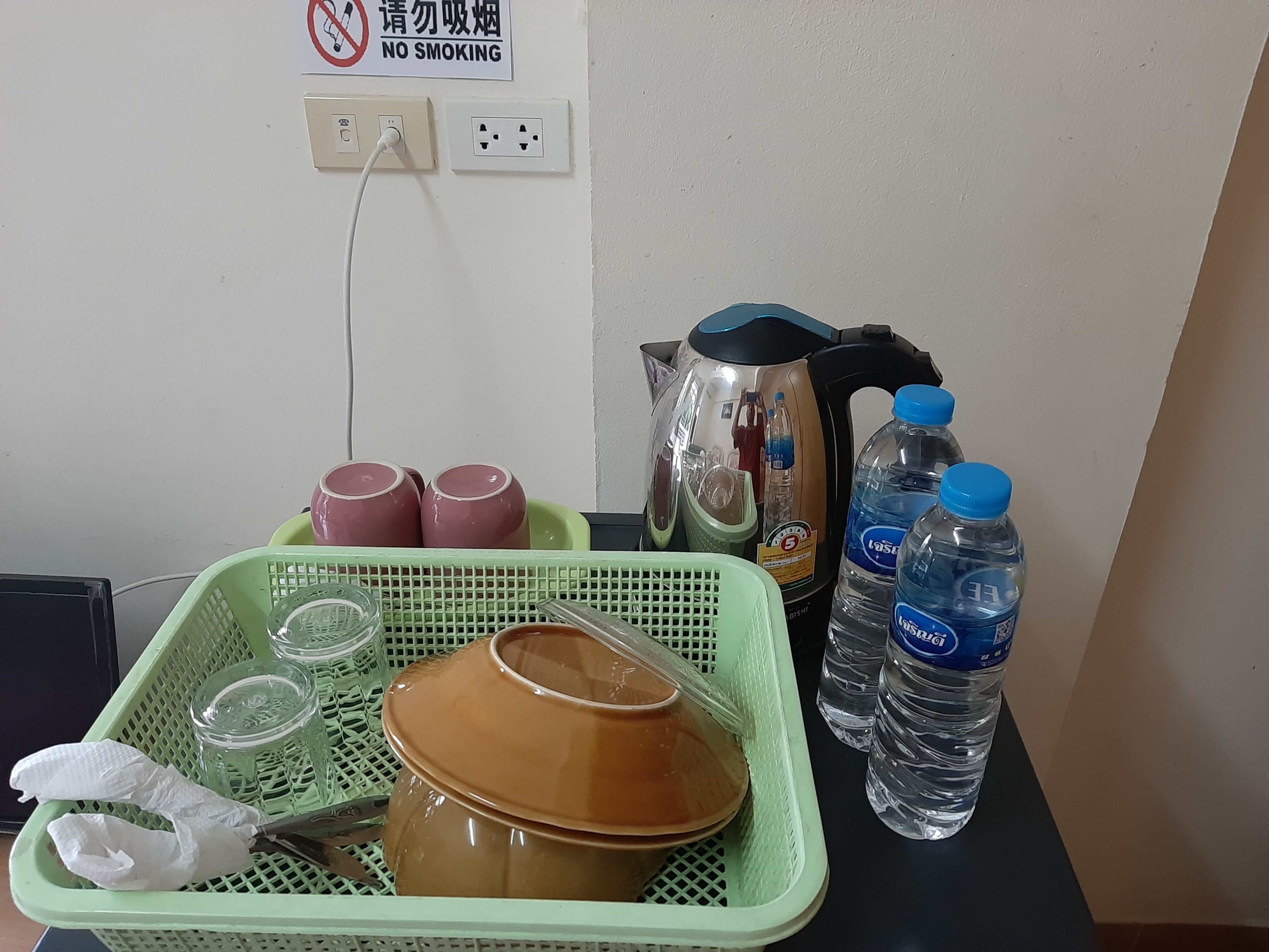 Una zona de cocina con un hervidor, botellas de agua, una cesta verde con utensilios de cocina y un cartel de 'No Smoking' en la pared.