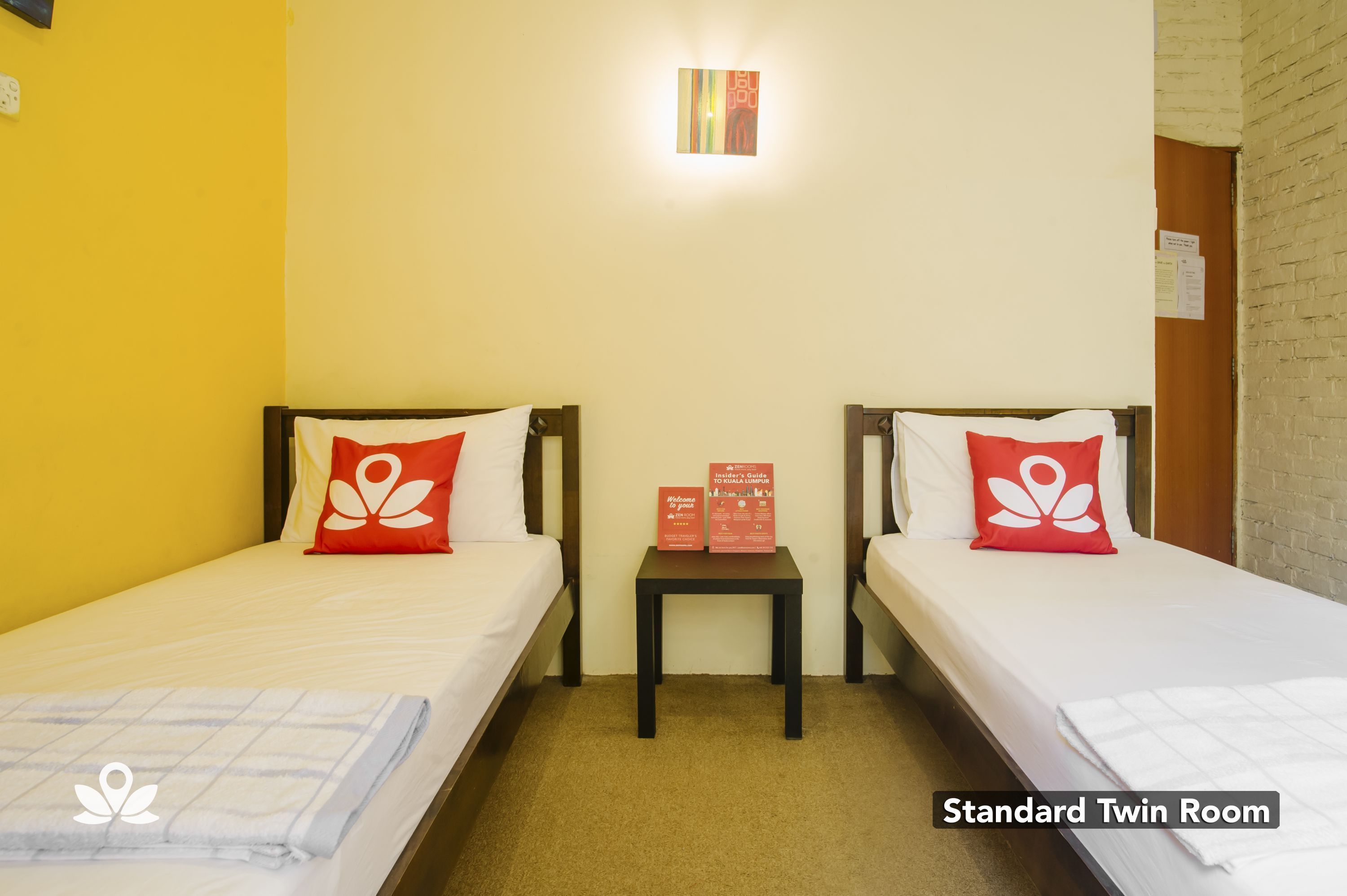 A standard twin room with two beds, each with a white mattress and a red pillow with a white flower design. A small table with a red brochure is between the beds.