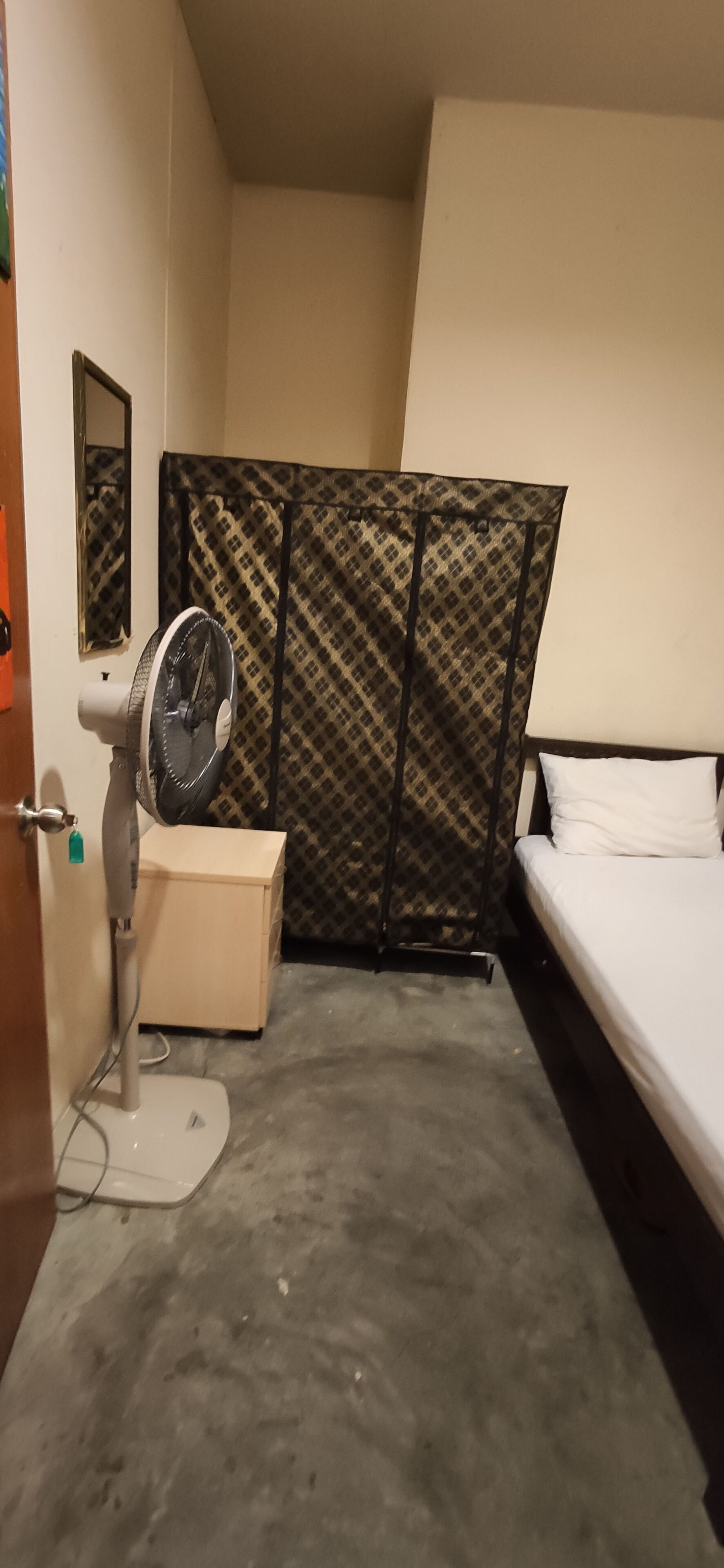 A small, compact room with a bed, a standing fan, a mirror, and a patterned curtain.