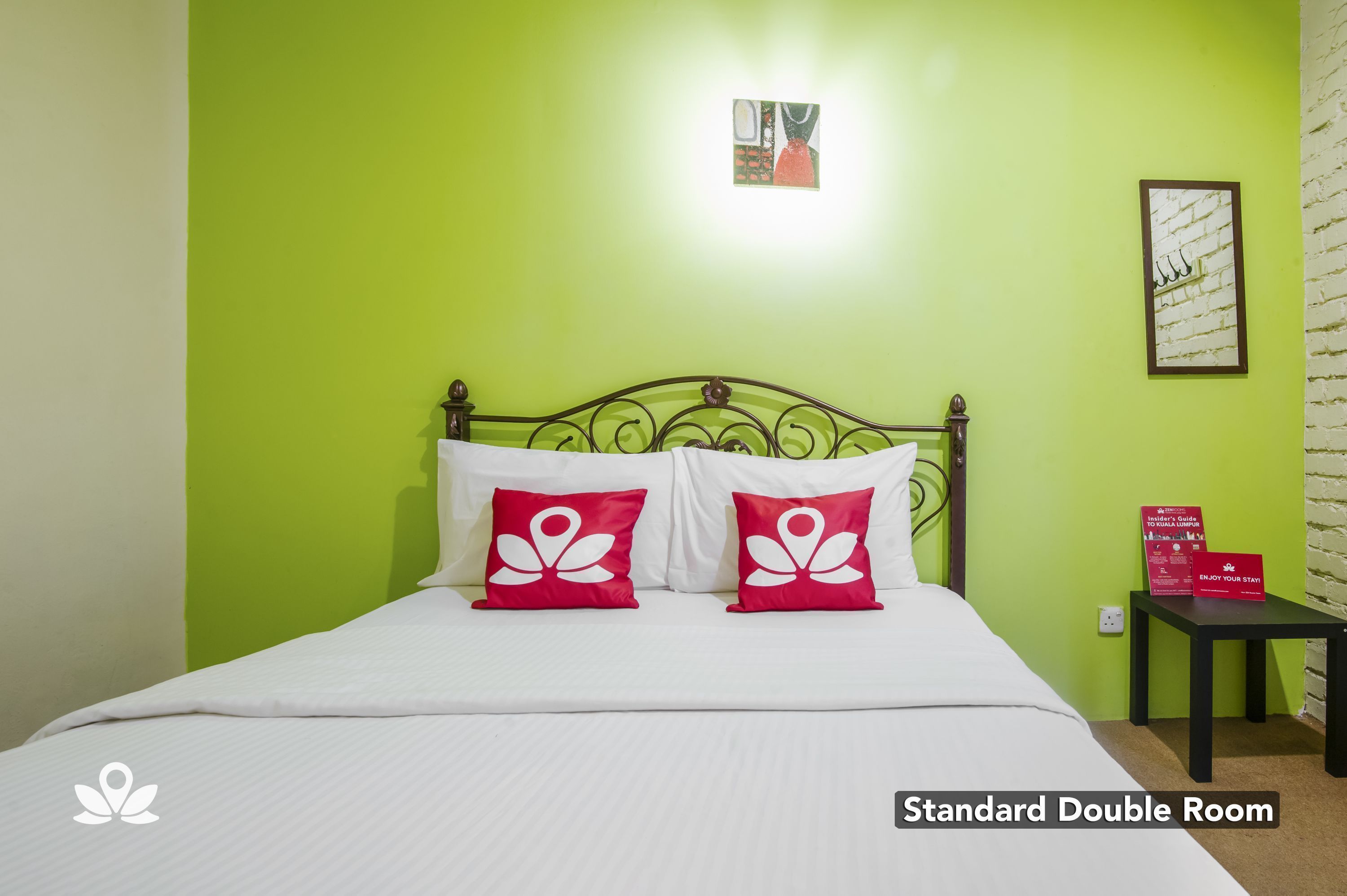 A standard double room with a bed, two red pillows, a nightstand, a framed picture, and a sign on the wall.