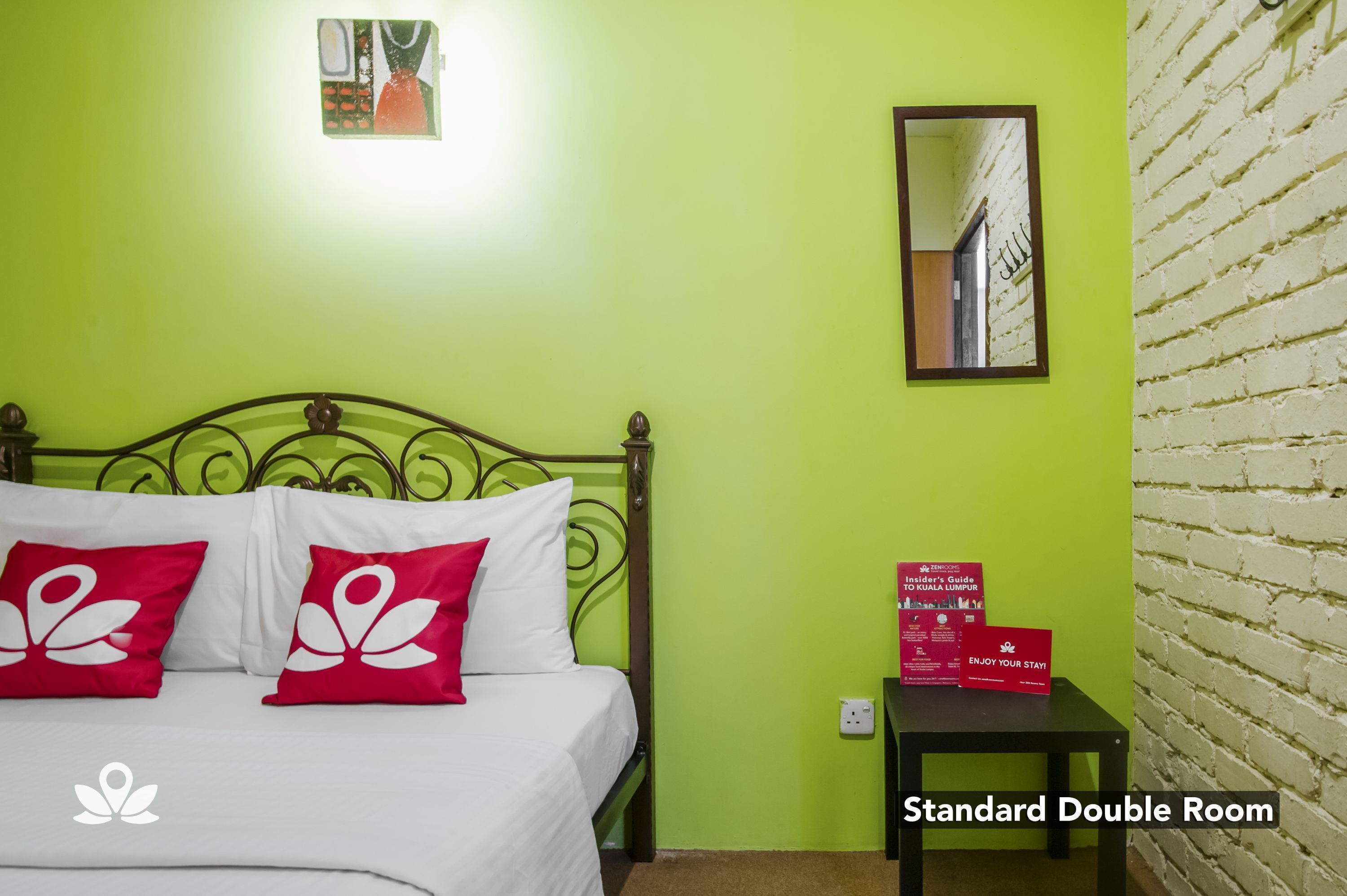 A standard double room with a green wall, a bed with white and red pillows, a small table, and a mirror.