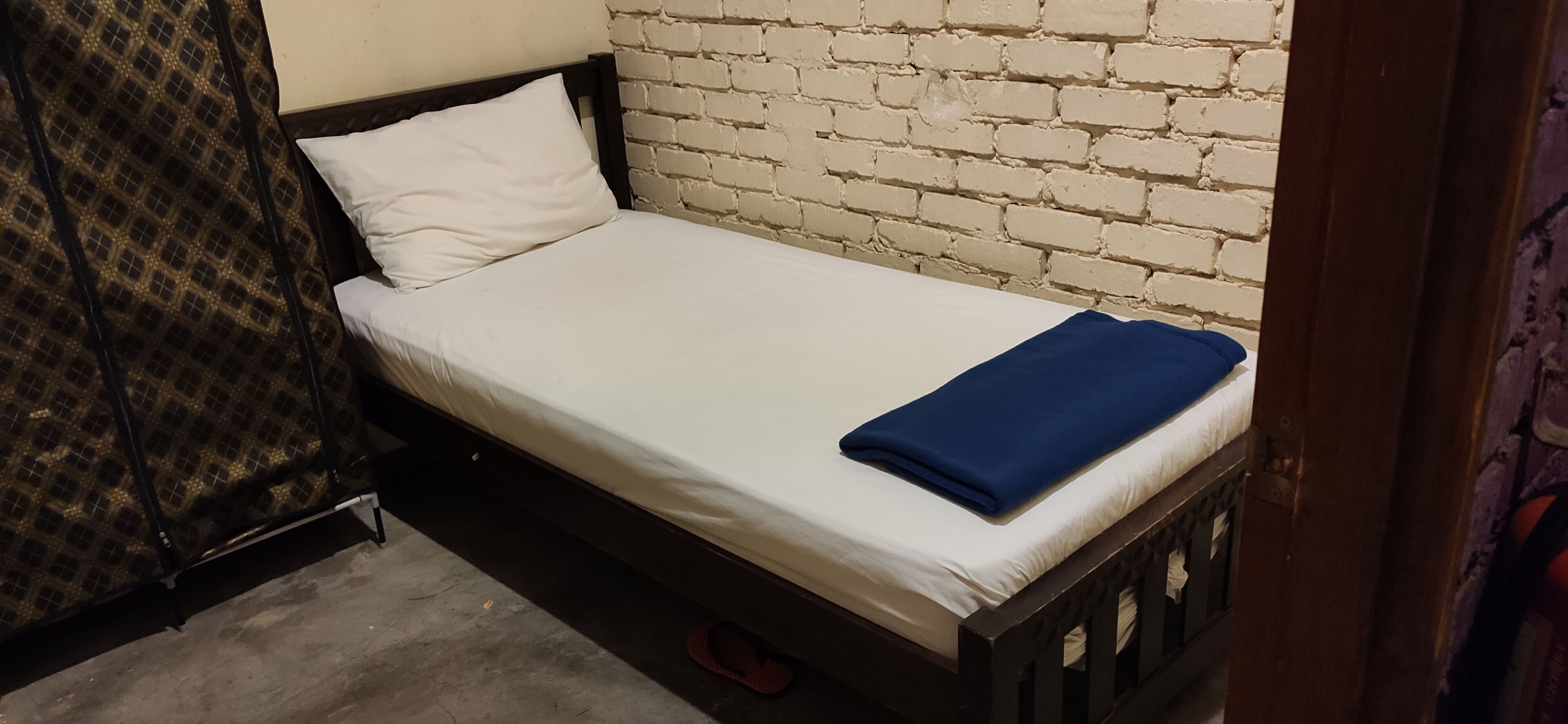 A small, single-bed room with a white mattress, a blue blanket, and a wooden headboard.