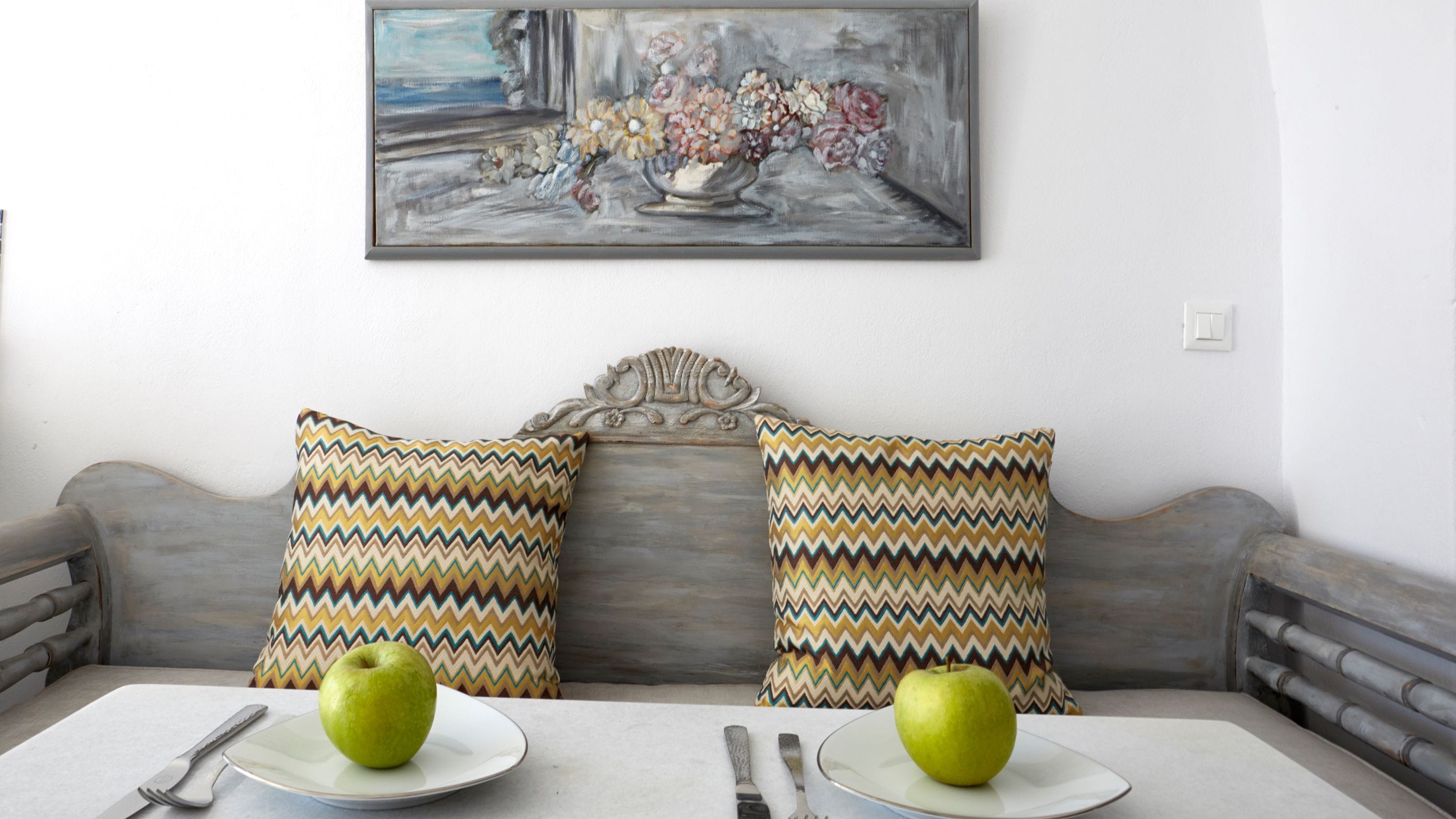 A dining area with a table set for two, featuring two green apples on white plates, a knife, and a fork. The table is in front of a grey sofa with two patterned pillows. Above the sofa, there is a framed painting of flowers.