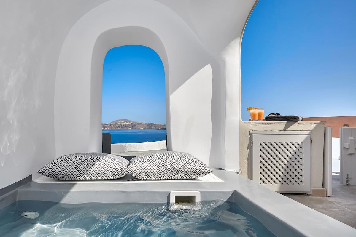 A small outdoor hot tub with two patterned pillows, a view of the sea, and a table with drinks and a towel.