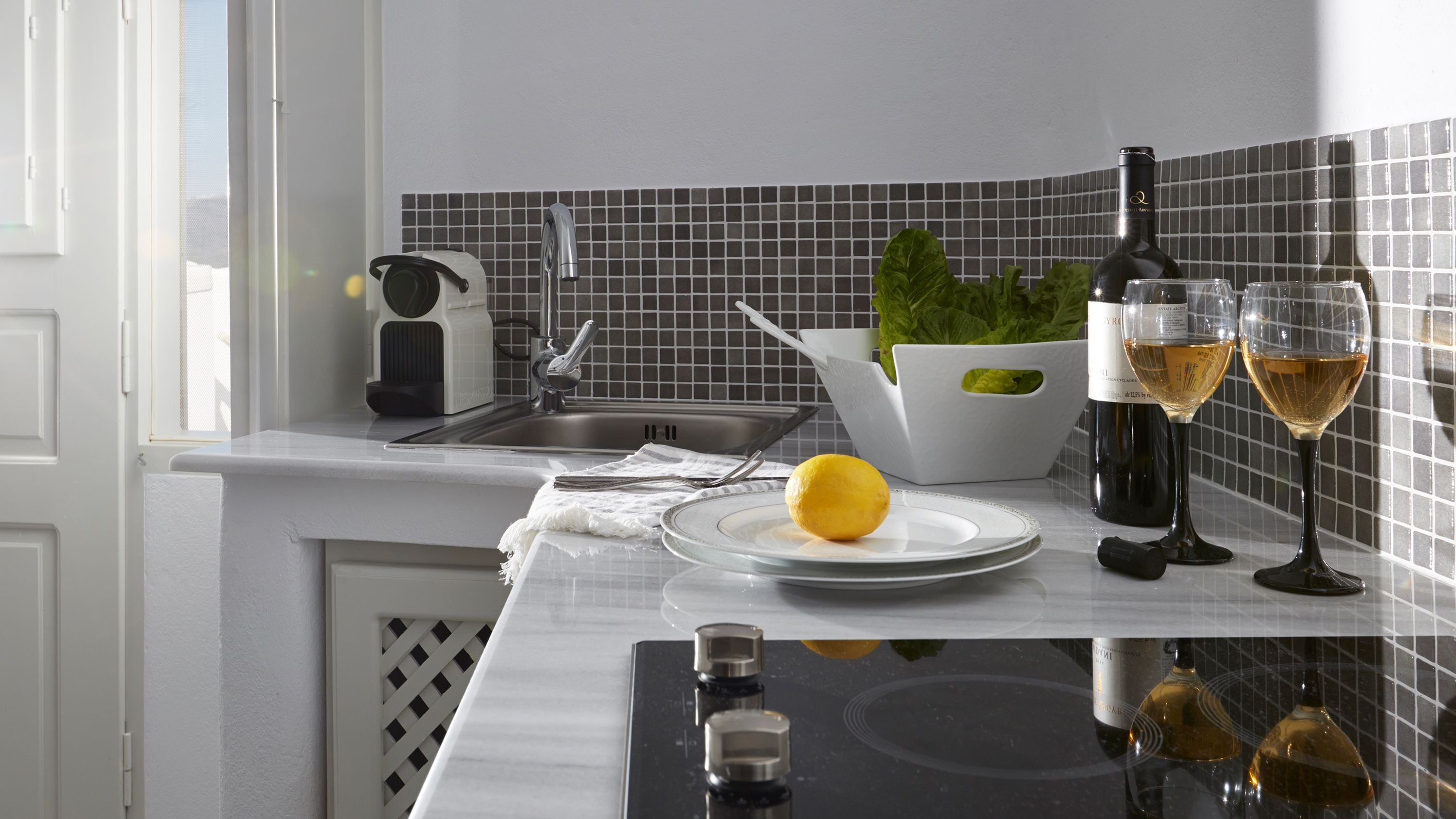 A kitchen with a sink, a white kettle, a white bowl, a potted plant, two wine glasses, and a lemon on a plate.