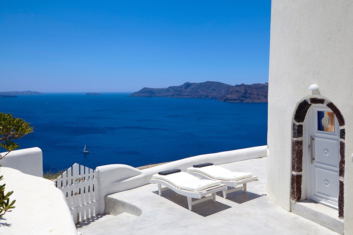 A white building with a door and a window, overlooking a blue sea with a sailboat.