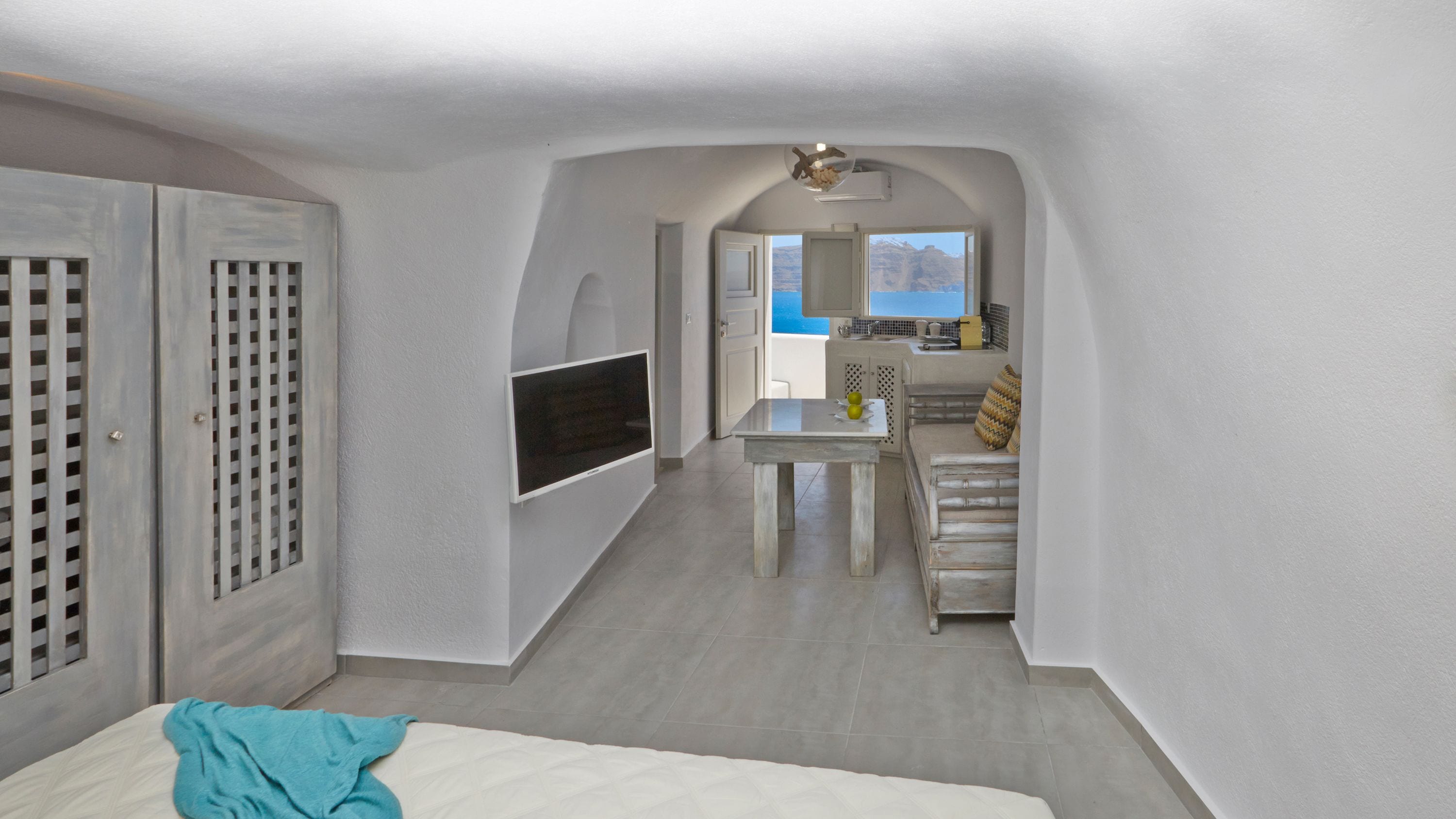 A compact hotel room with a bed, a TV mounted on the wall, a kitchenette, and a view of the mountains.