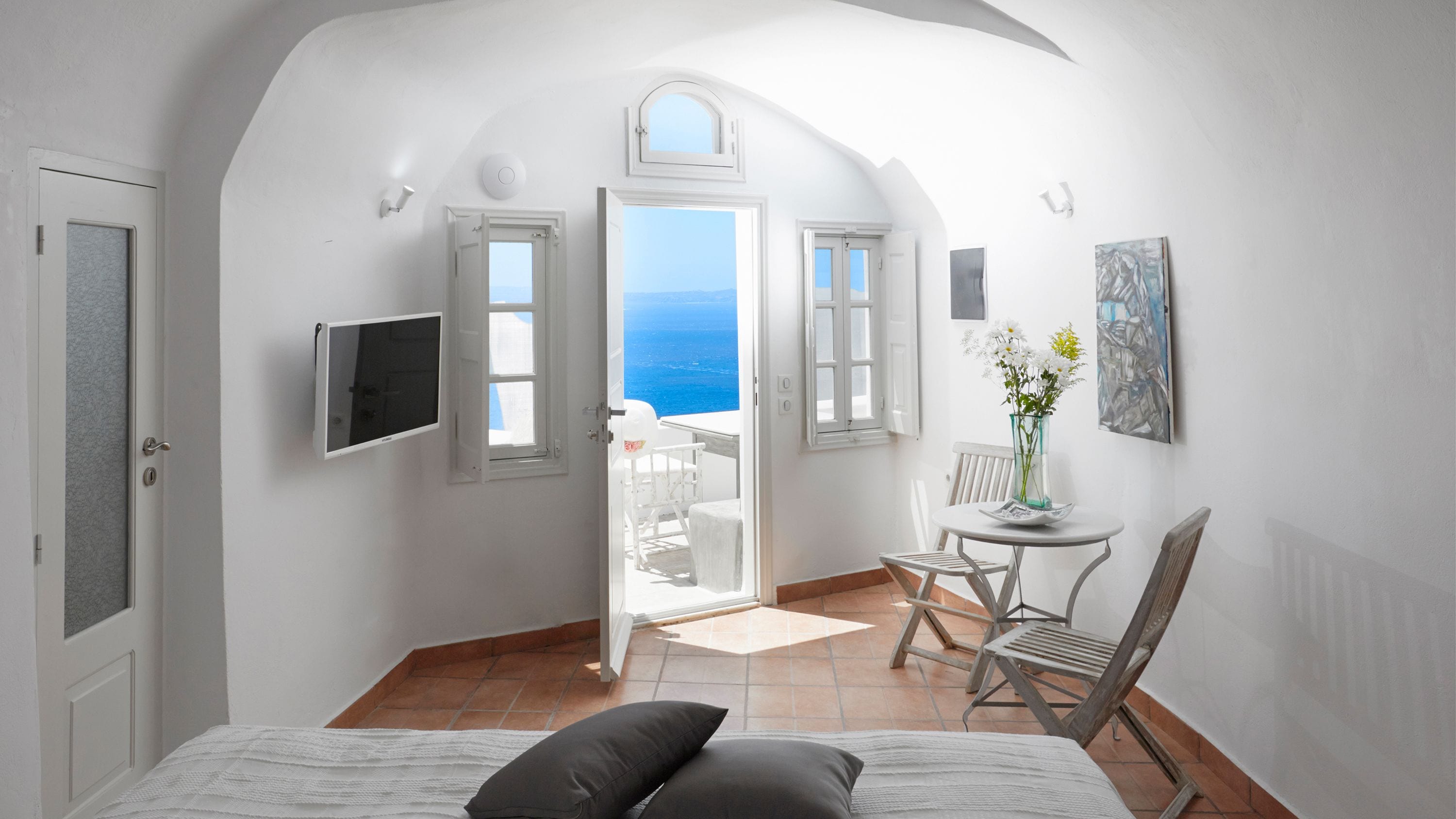 A bedroom with a bed, a chair, a small table, a TV, and a view of the sea.