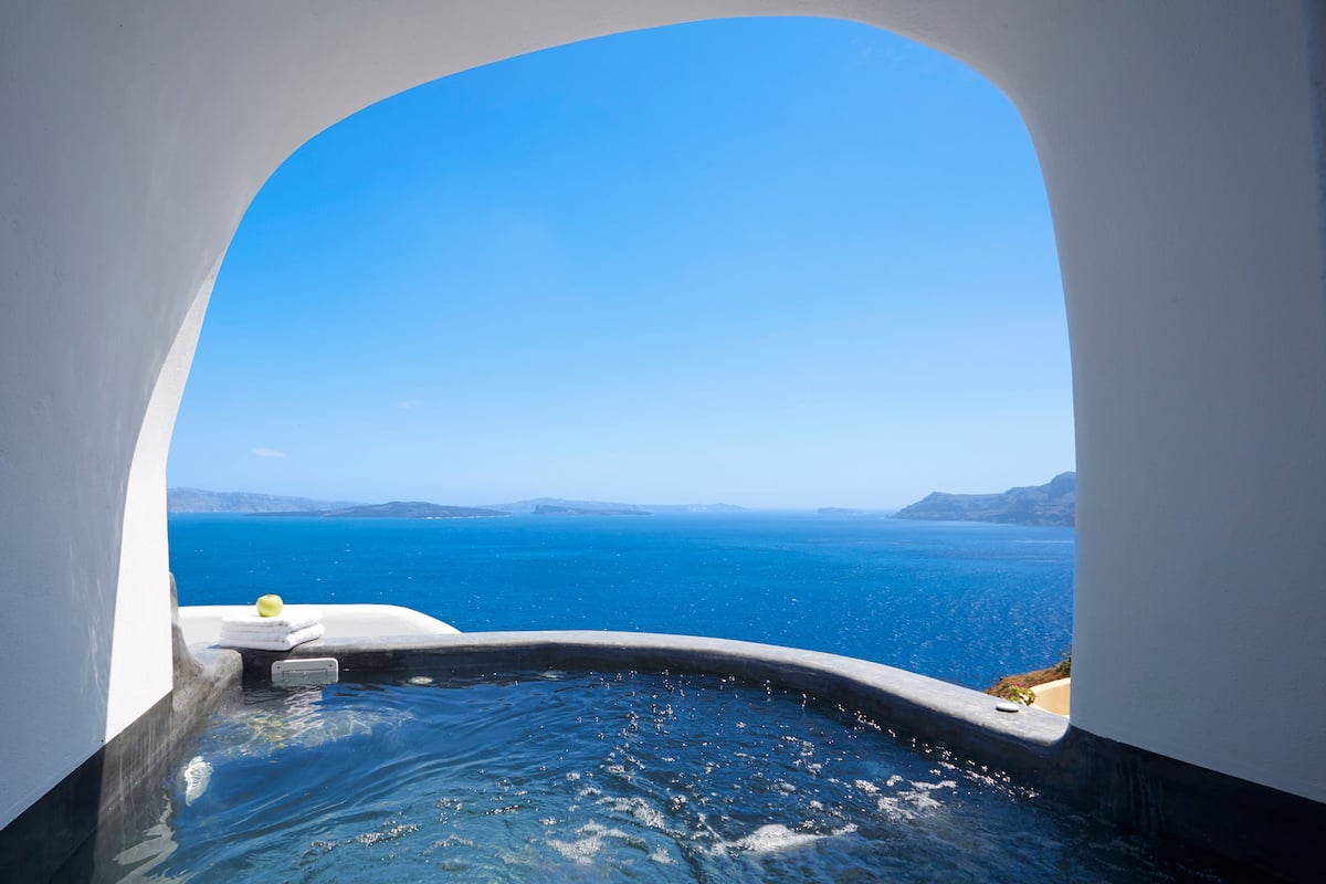 A view through a white arch window of a pool with clear blue water, overlooking a vast ocean and distant land under a bright blue sky.