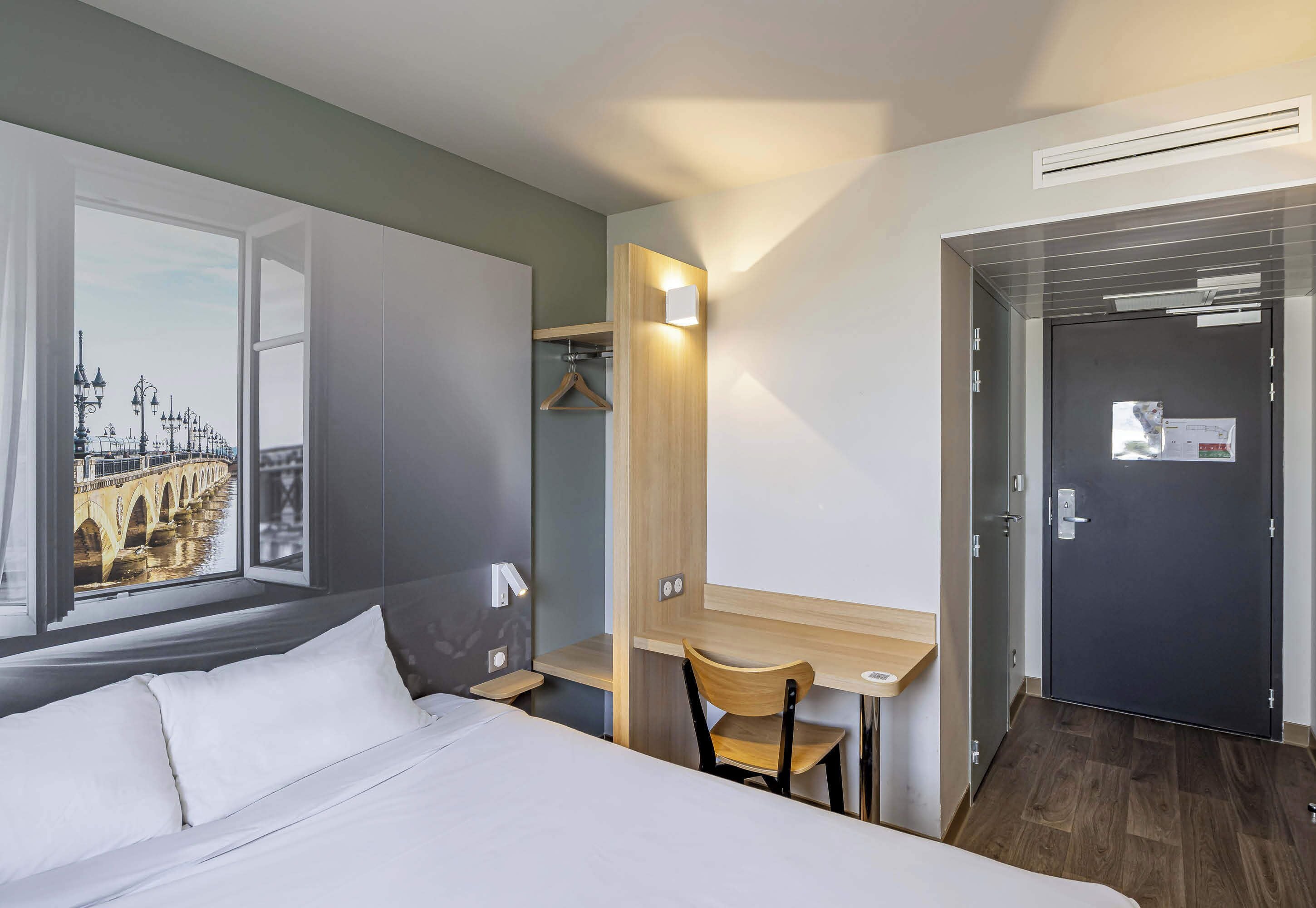 A hotel room with a bed, a desk, a chair, a wardrobe, and a door.