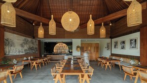 Uma ampla área de refeições com longas mesas e cadeiras de madeira, luminárias pendentes trançadas e um grande mural na parede.